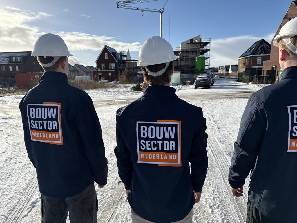 Drie vakmensen van Bouwsector Nederland in blauwe werkjassen en witte helm tussen nieuwe huizen